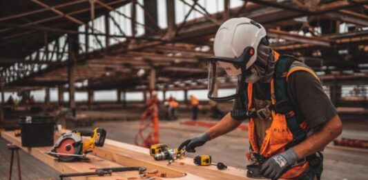 Why Builders Need Public Liability Insurance man in orange and black vest wearing white helmet holding yellow and black power tool