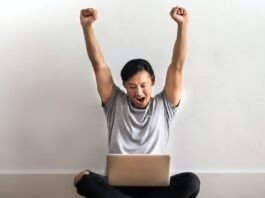 Blue Dragon Casino: Top 3 Exceptional Games to Play! Photo of Man in Gray T-shirt and Black Pants Sitting on Wooden Floor and Working on His Laptop Celebrating with His Hands Raised