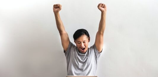 Blue Dragon Casino: Top 3 Exceptional Games to Play! Photo of Man in Gray T-shirt and Black Pants Sitting on Wooden Floor and Working on His Laptop Celebrating with His Hands Raised