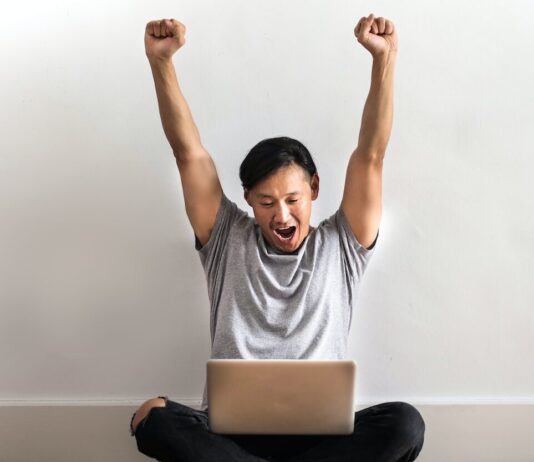 Blue Dragon Casino: Top 3 Exceptional Games to Play! Photo of Man in Gray T-shirt and Black Pants Sitting on Wooden Floor and Working on His Laptop Celebrating with His Hands Raised