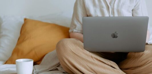 The Rise of Online Casino Entertainment in Singapore: A Tale of Digital Euphoria Crop faceless female in casual outfit sitting on bed with legs crossed holding laptop on knees with cup of coffee standing on notebook while working from home