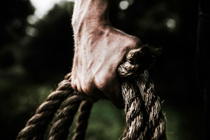 Photo by Evelyn Chong A close-up image shows a strong, firm hand gripping a thick, rough rope, showcasing strength and determination in an outdoor setting.