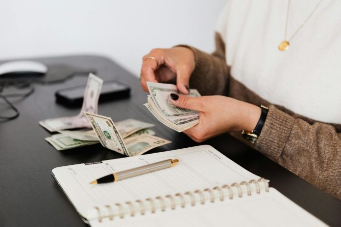 Photo by Photo By: Kaboompics.com Close-up of person counting cash with notepad on desk, indicating financial tasks.