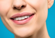The Morning Habit That’s Quietly Damaging Your Teeth A close-up image of a woman's smile with pink lipstick and white teeth against a blue background.