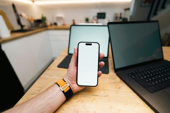Photo by Jakub Żerdzicki A hand holds a phone with two laptops behind it.